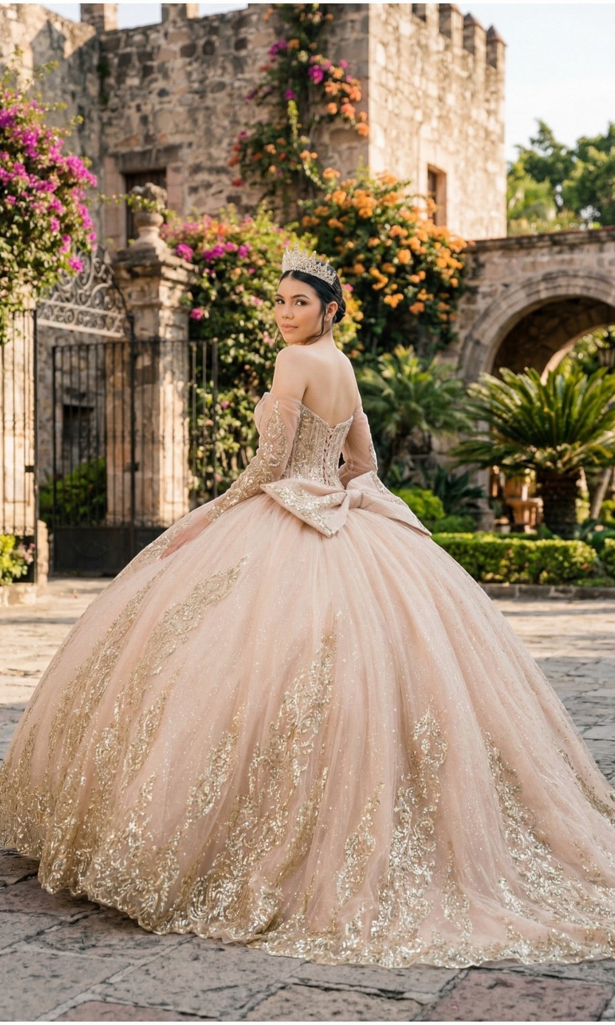 A young woman wearing the Quinceanera Dress 8545JQ by Cinderella Couture—featuring pink and gold beaded sequins and an off-the-shoulder sweetheart neckline—stands outdoors before a stone building and flowering plants, glancing over her shoulder.